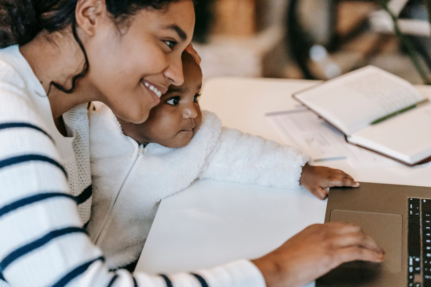 A mother smiling with her baby close to her face while working at a laptop, notebook open beside them.