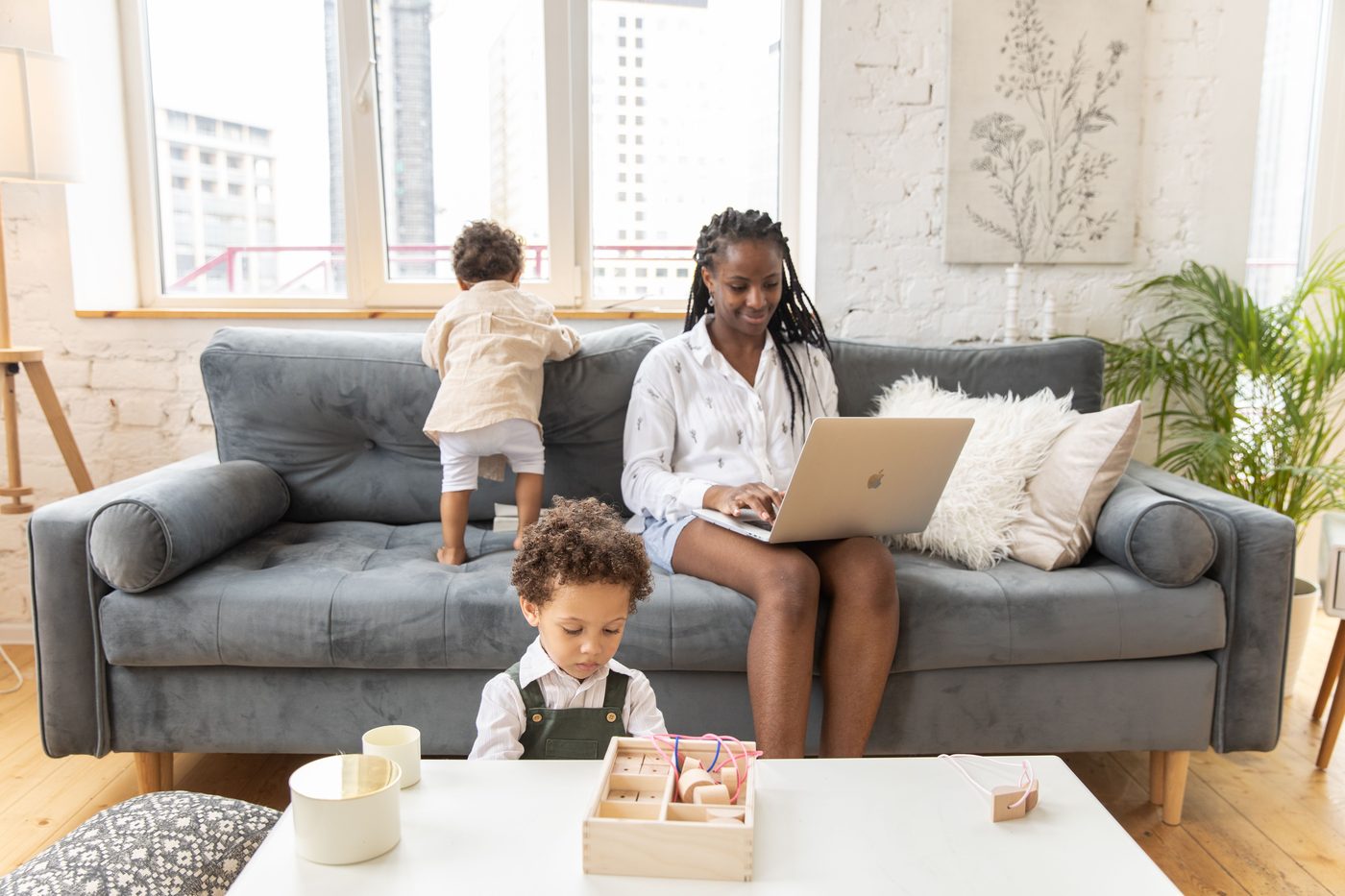 A mother working on her laptop on the sofa while her two young children play and explore around her.
